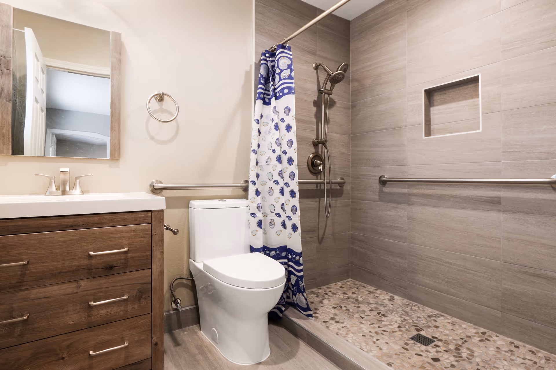 Accessible bathroom with a wooden vanity, toilet, and a walk-in tiled shower with grab bars and a blue patterned shower curtain.