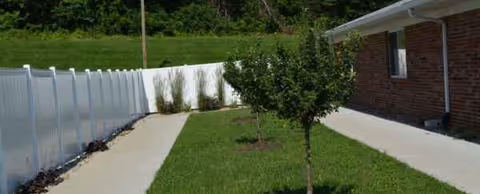 Side of brick facility with a concrete walkway, white fence, and small trees on lawn.