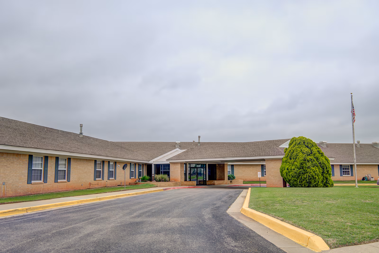 Single-story brick assisted living facility with a covered central entrance, driveway, lawn, and flagpole under a cloudy sky.