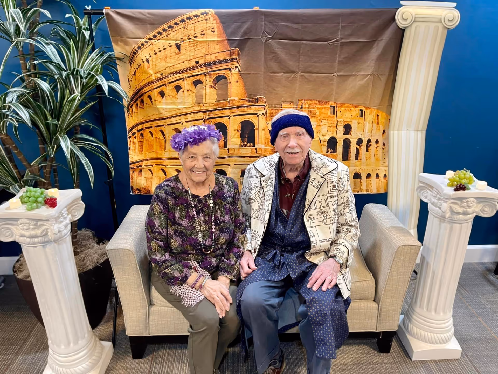 An elderly woman and man sitting on a beige couch in front of a backdrop featuring the Colosseum. The woman is wearing a purple floral top and a purple flower crown, while the man is dressed in a patterned jacket and a dark blue hat. They are flanked by two white decorative columns topped with grapes and cheese, and a potted plant is visible on the left side.