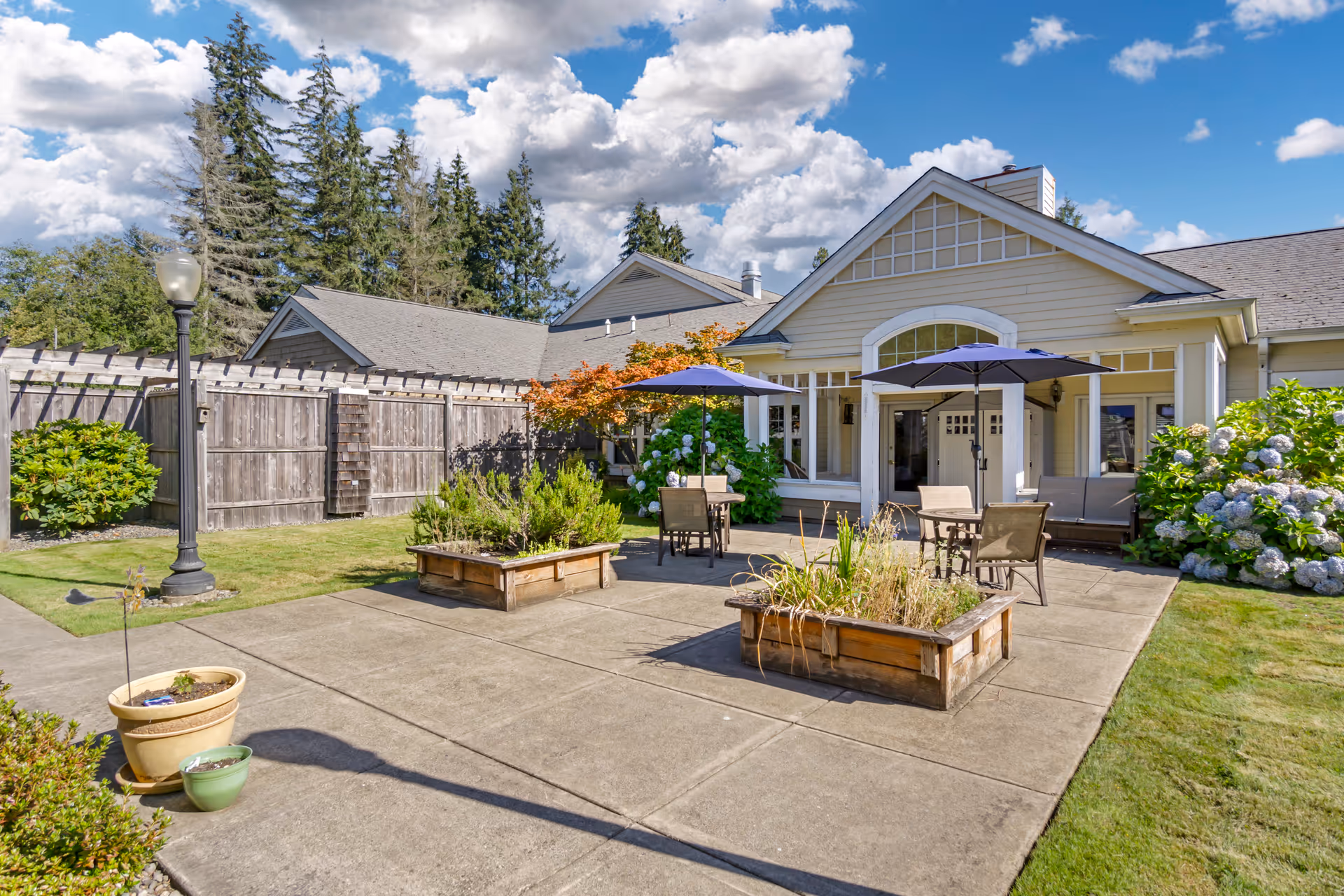 Outdoor patio area at Brookdale Olympia West with tables, chairs, and umbrellas on a concrete surface surrounded by greenery, flower beds, and a wooden fence under a partly cloudy sky.
