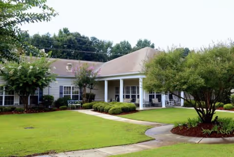 Single-story building with a covered porch entrance surrounded by well-maintained green lawn, shrubs, and trees under a partly cloudy sky.