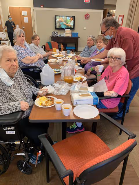 A group of elderly people sitting around a dining table in a senior living facility, enjoying a meal together. There are plates with food, cups with beverages, and a pitcher on the table. One man is standing and interacting with a woman in a pink shirt. A television is visible in the background, along with some furniture and a staff member wearing a mask.