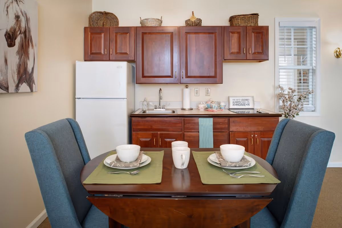 Small kitchenette with dark wood cabinets, a white refrigerator and sink, and a dining table set for two with teal chairs.