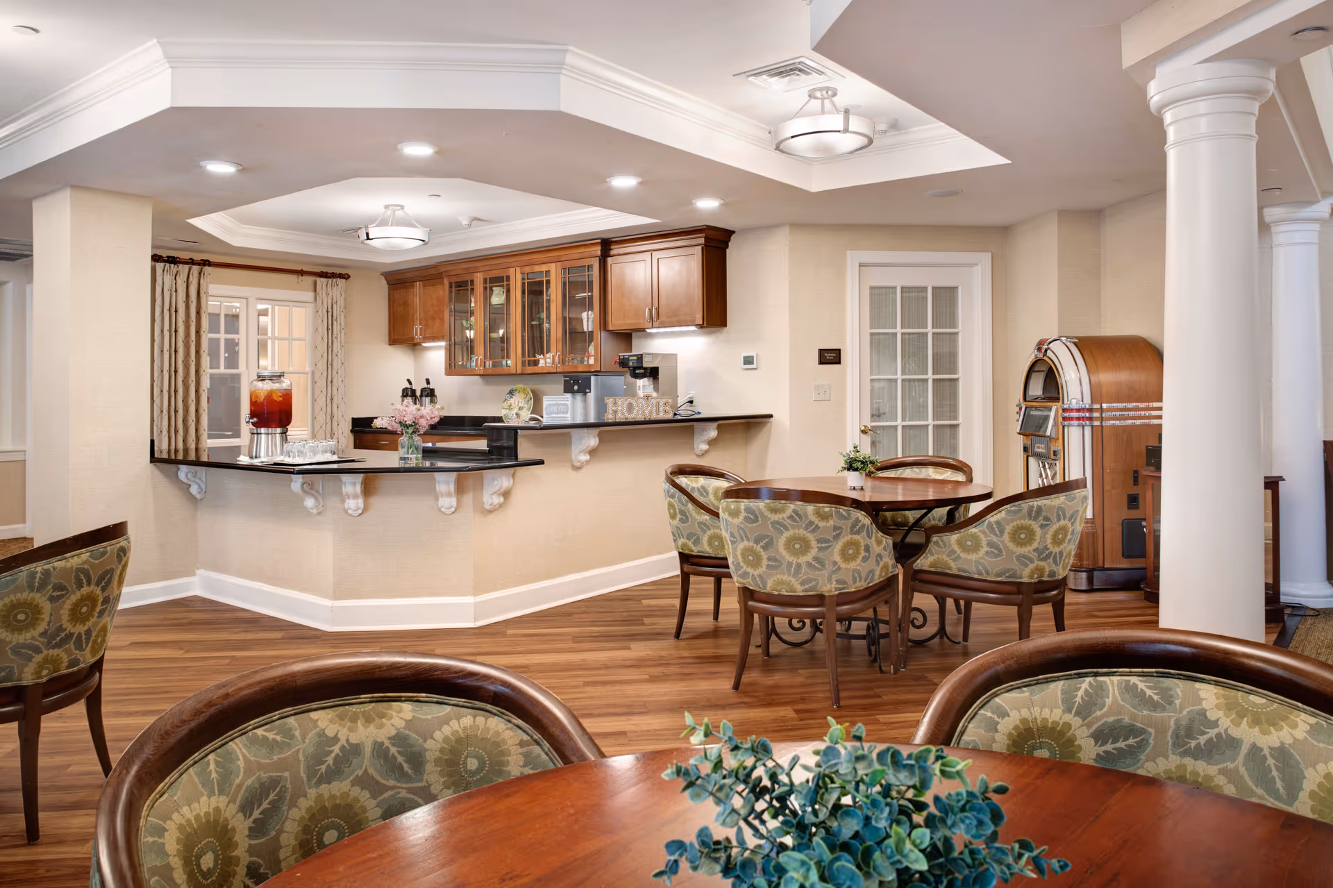 Warm communal dining and lounge area with round tables and upholstered chairs, a serving counter with cabinets, and a jukebox.