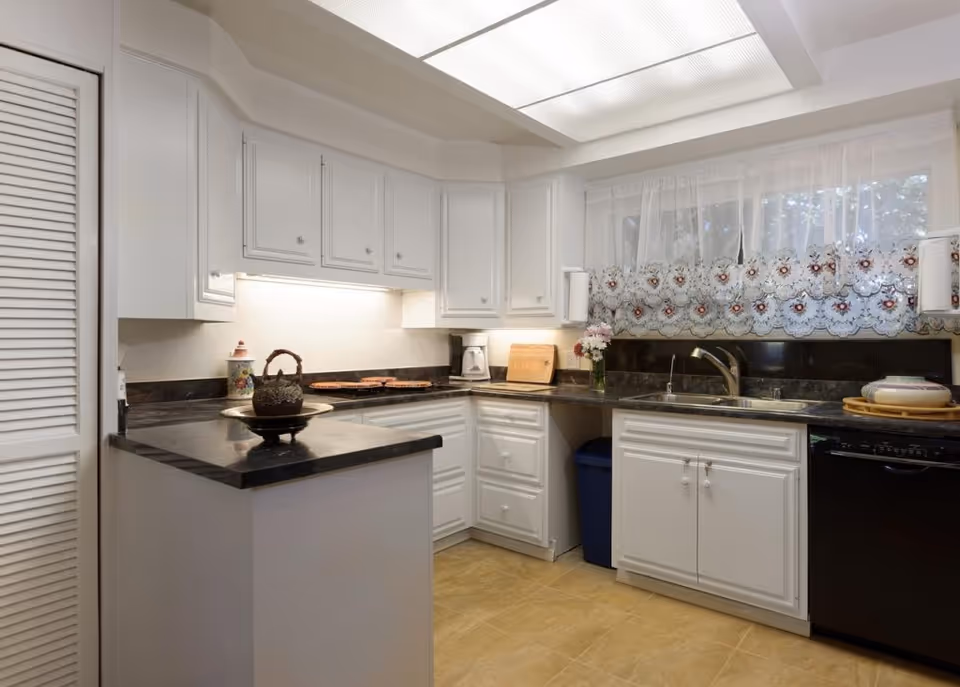 Bright kitchen with white cabinets, dark countertops, a sink under a lace-curtained window, and a small peninsula island.