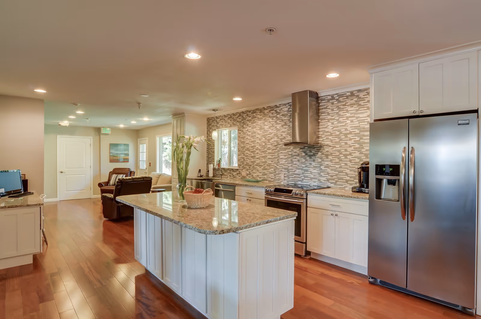 Modern kitchen with white cabinets, a large island with granite countertop, stainless steel refrigerator, stove, and range hood. The kitchen opens into a living area with leather chairs and a sofa, hardwood floors, and recessed lighting.