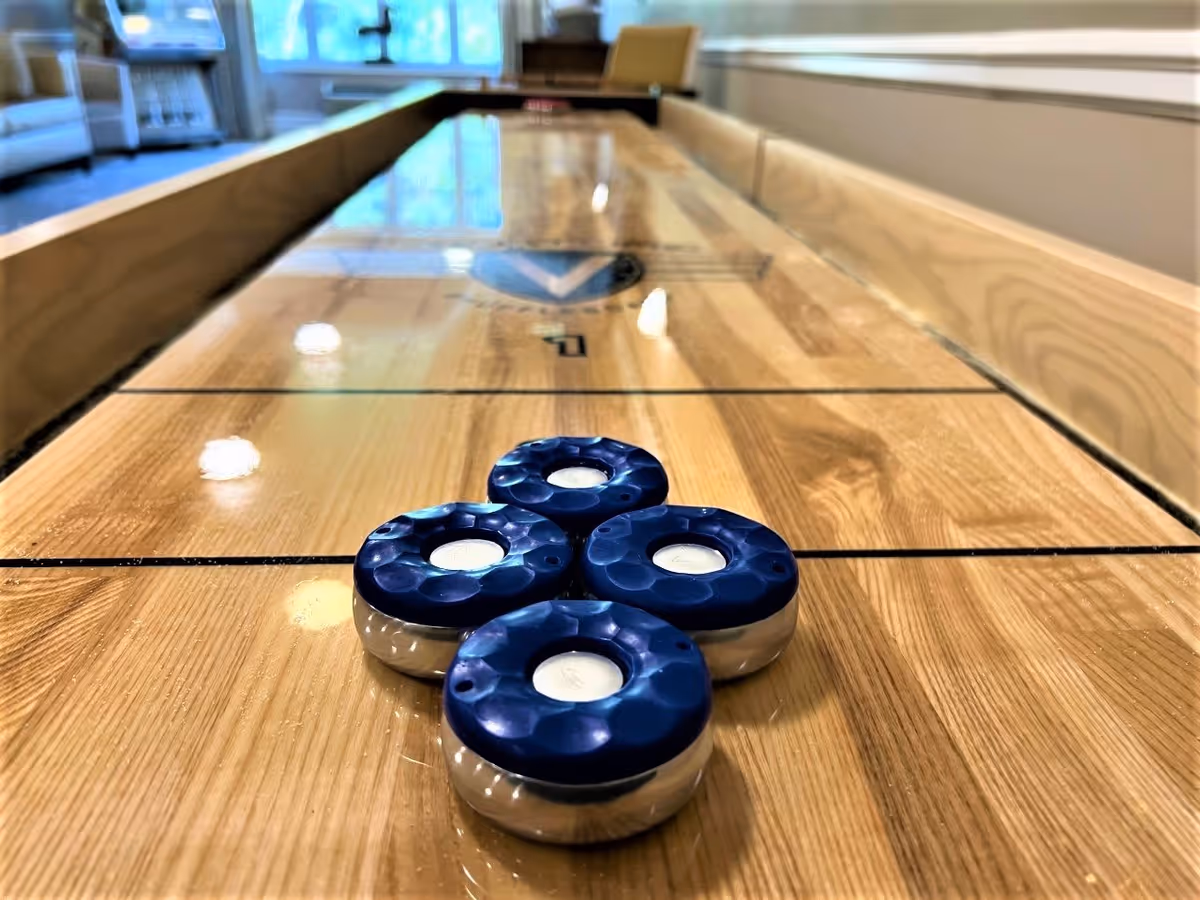 Close-up view of a shuffleboard table with four blue pucks grouped together on the wooden playing surface in a well-lit room with windows and seating in the background.