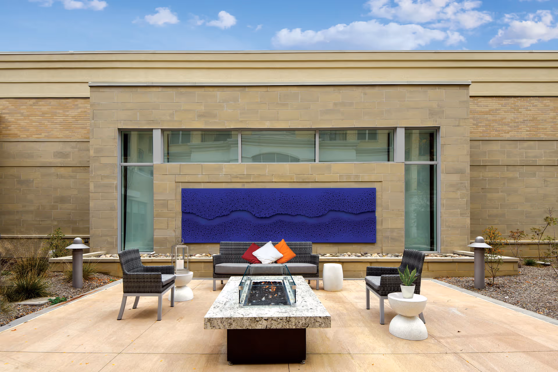 Outdoor patio area with modern wicker seating including a sofa and two chairs arranged around a rectangular stone fire pit. The sofa has three colorful cushions in red, white, and orange. A small white side table with a potted plant is next to one chair. The background features a beige brick wall with tall windows and a large blue abstract wall art piece mounted on it. The sky is partly cloudy.