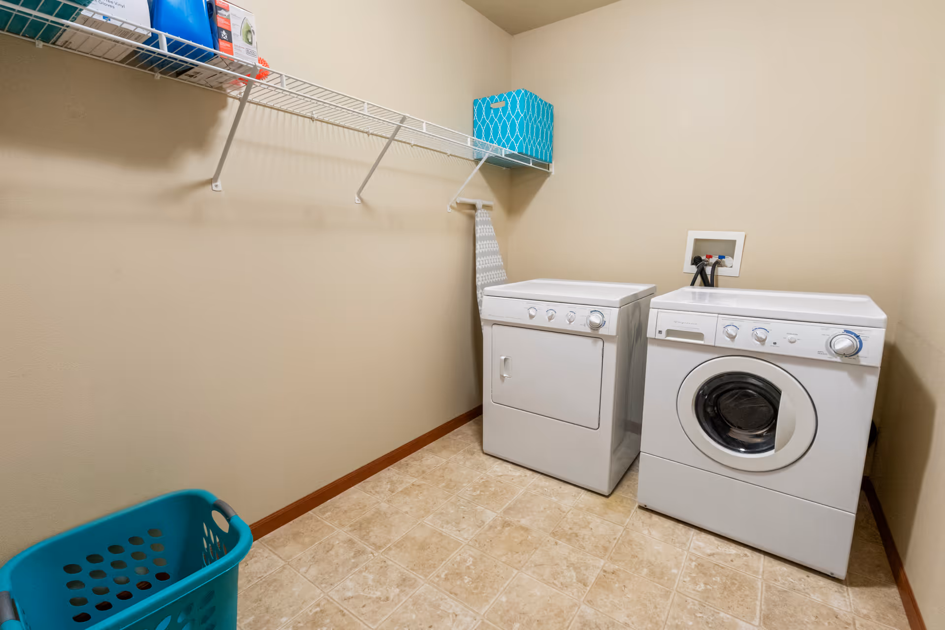 Laundry room with a white washing machine and dryer side by side against a beige wall. Above them is a white wire shelf holding a blue patterned storage box and some laundry supplies. A turquoise laundry basket is on the tiled floor in the corner.