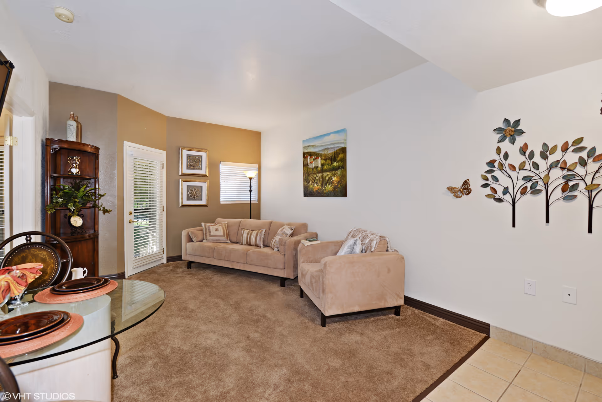 A cozy living room with beige carpet and light-colored walls. The room features a beige sofa and matching armchair with decorative pillows. There is a floor lamp near a window with blinds, a glass-top dining table with place settings, and a wooden corner shelf with plants and decorative items. Wall decorations include framed artwork and metal tree and butterfly sculptures.