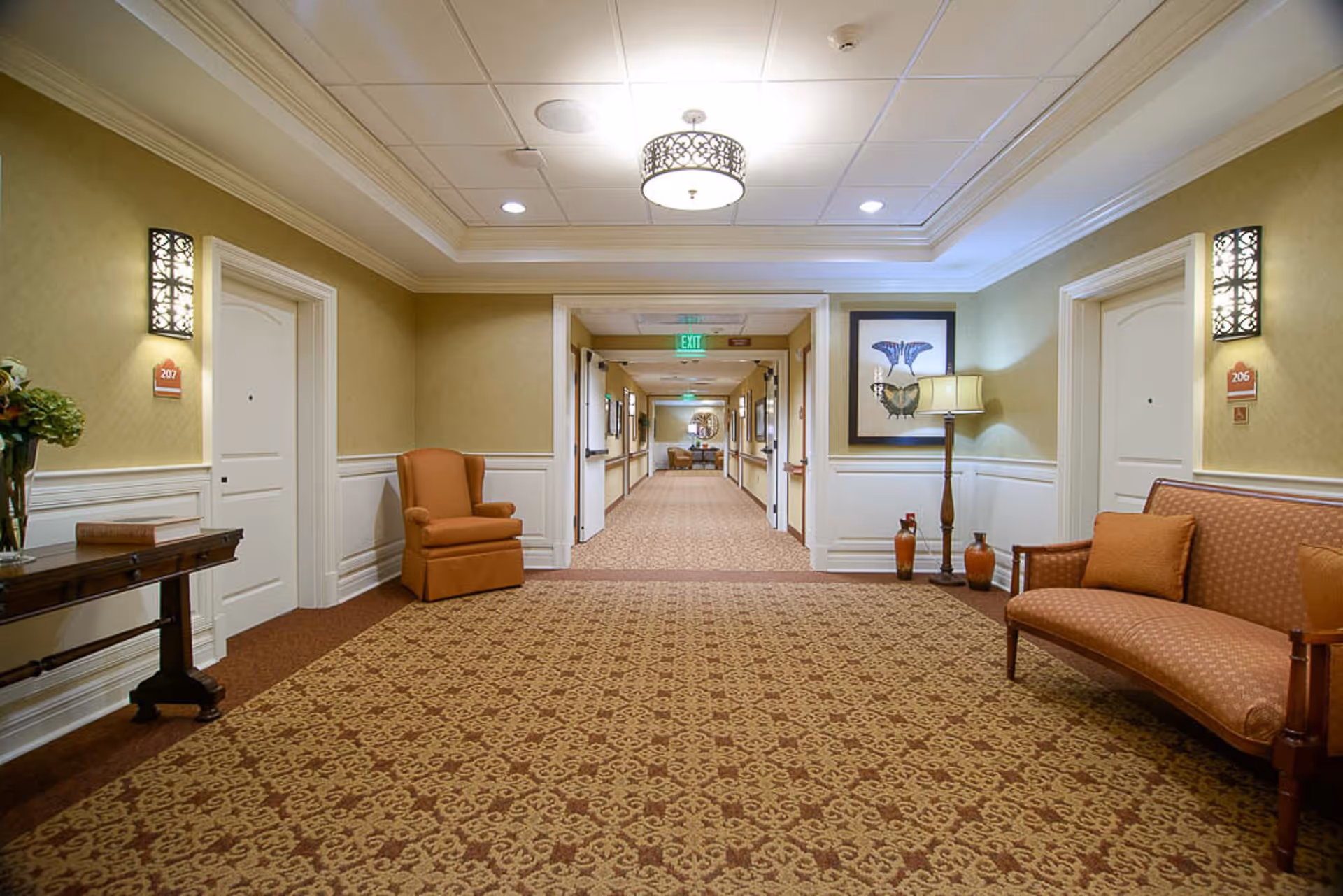 A well-lit hallway in a senior living facility with patterned carpet and beige walls. There are two white doors on either side labeled 206 and 207. The hallway is furnished with an orange armchair, a matching couch with cushions, a wooden side table with a vase of flowers, a floor lamp, and decorative wall sconces. Artwork and decorative vases are also visible along the walls.