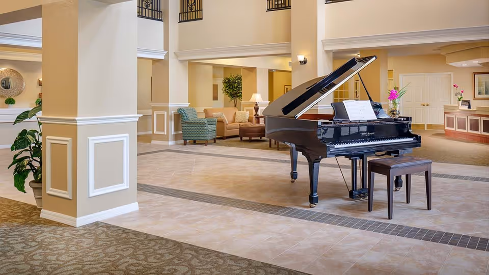 Spacious and well-lit assisted living facility common area featuring a black grand piano with sheet music on the stand, a wooden piano bench, beige tiled flooring with a decorative border, and comfortable seating including a green patterned armchair and a beige sofa. The area is decorated with plants, wall sconces, and a reception desk in the background.