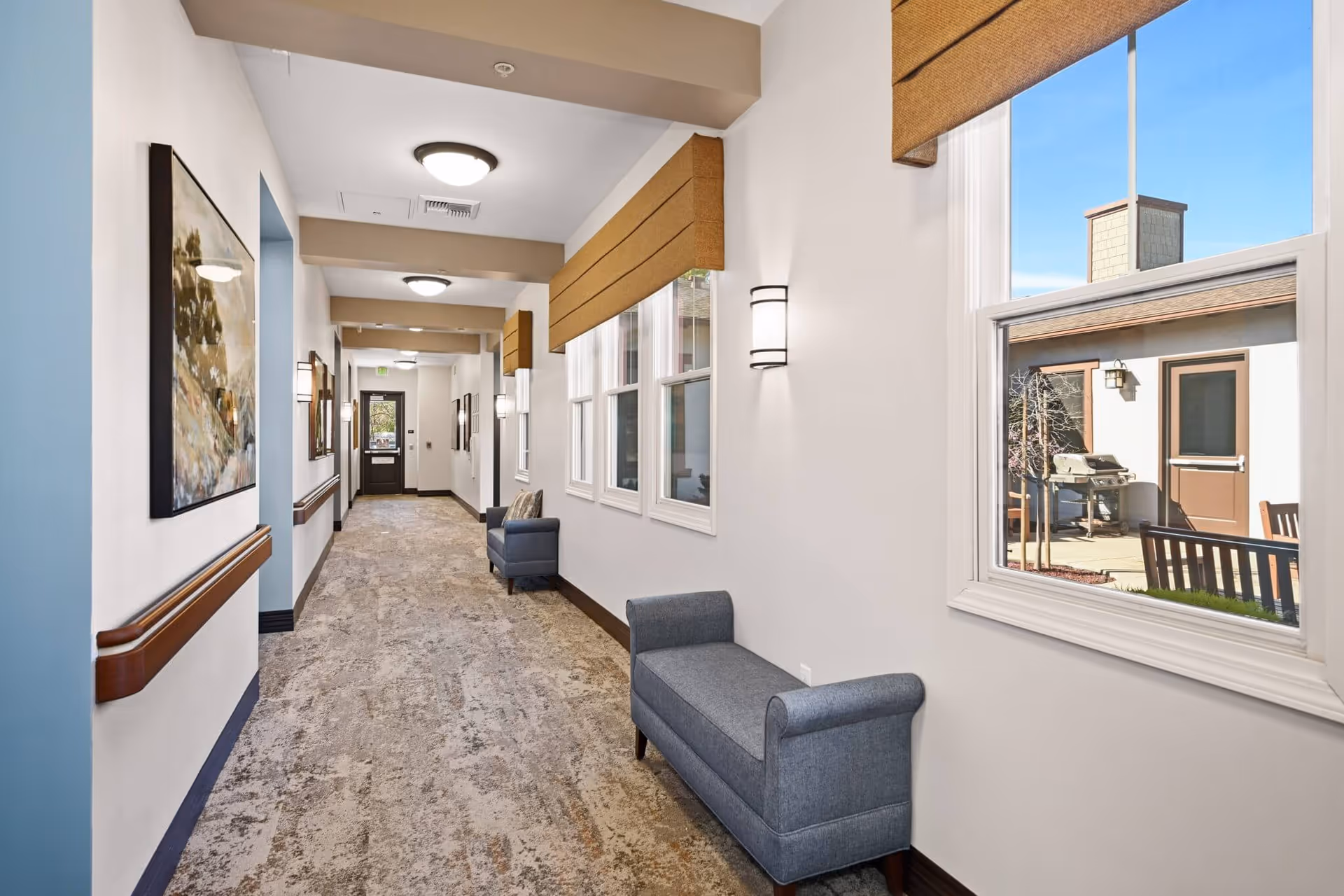 A well-lit hallway in a senior living facility with beige walls, carpeted floor, and ceiling lights. There are two blue upholstered benches along the right wall beneath windows with brown valances. The left wall features a large framed painting and wooden handrails. Outside the window, a patio area with a barbecue grill and outdoor furniture is visible.