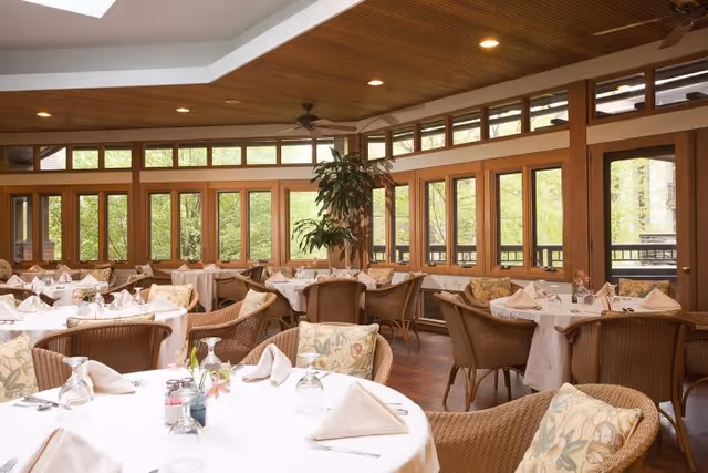 A bright and spacious dining room with multiple round tables covered in white tablecloths, each set with folded napkins, glasses, and cutlery. The room features large windows with wooden frames allowing natural light to fill the space, wicker chairs with floral cushions, a wooden ceiling with recessed lighting, and ceiling fans. There are plants near the windows adding a touch of greenery.