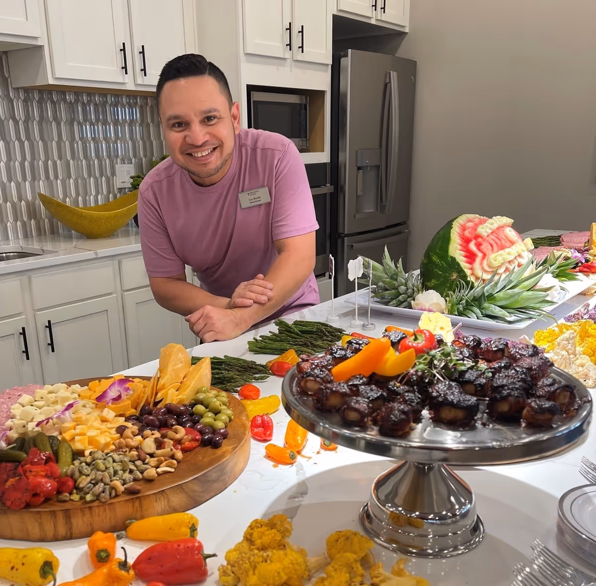 A smiling man wearing a pink shirt and a name tag stands behind a kitchen counter filled with an assortment of food including a cheese and nut platter, grilled asparagus, stuffed dates on a silver pedestal, and a decorative watermelon carving. The kitchen has white cabinets and a stainless steel refrigerator in the background.