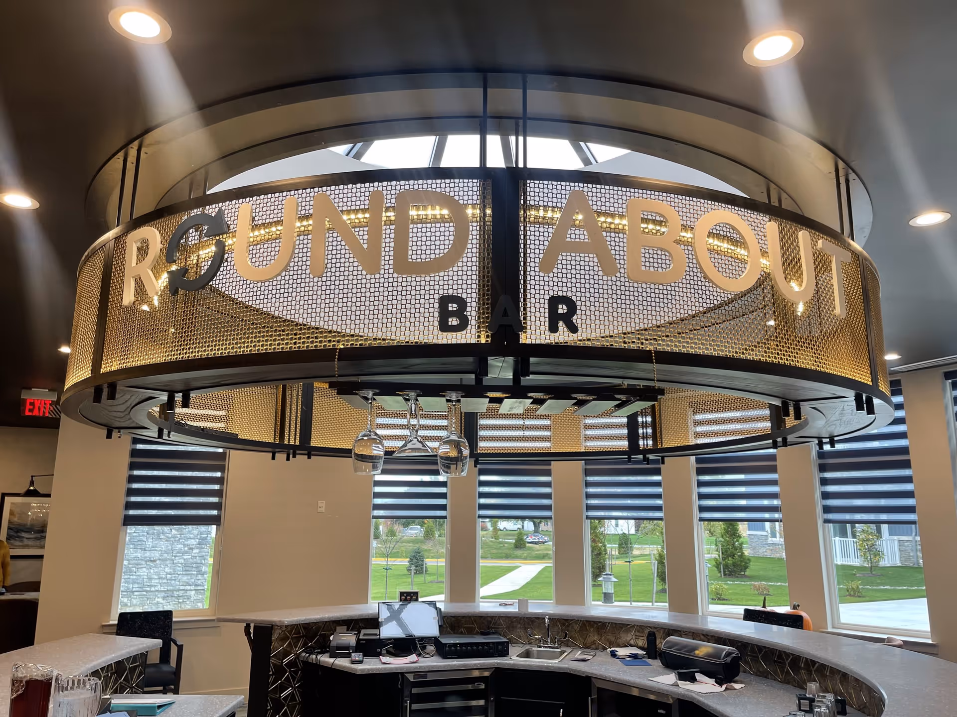Interior view of a circular bar area named 'Round About Bar' with a curved counter, hanging wine glasses, and large windows with striped blinds showing a green outdoor area.