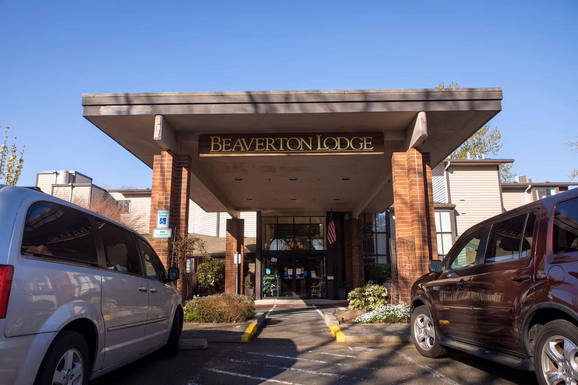 Entrance of Beaverton Lodge senior living facility with a covered drop-off area supported by brick pillars, two parked vehicles on either side, and clear blue sky above.