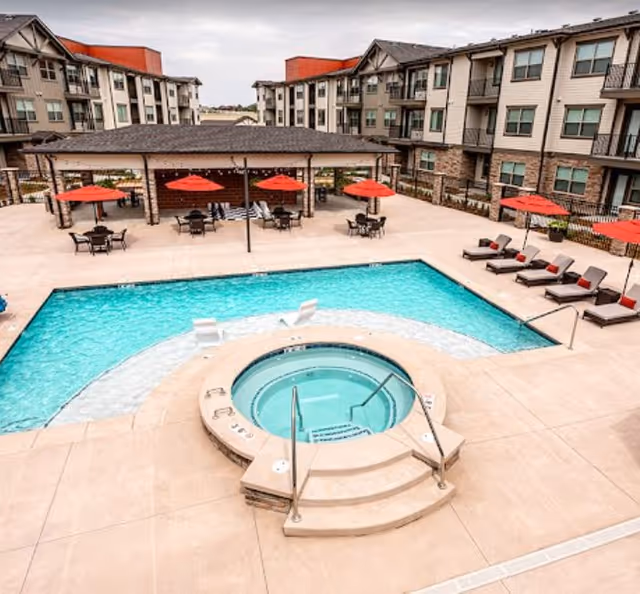 Outdoor swimming pool area with a circular hot tub in the foreground, surrounded by lounge chairs with red cushions and red umbrellas. The pool area is enclosed by a multi-story residential building with balconies.