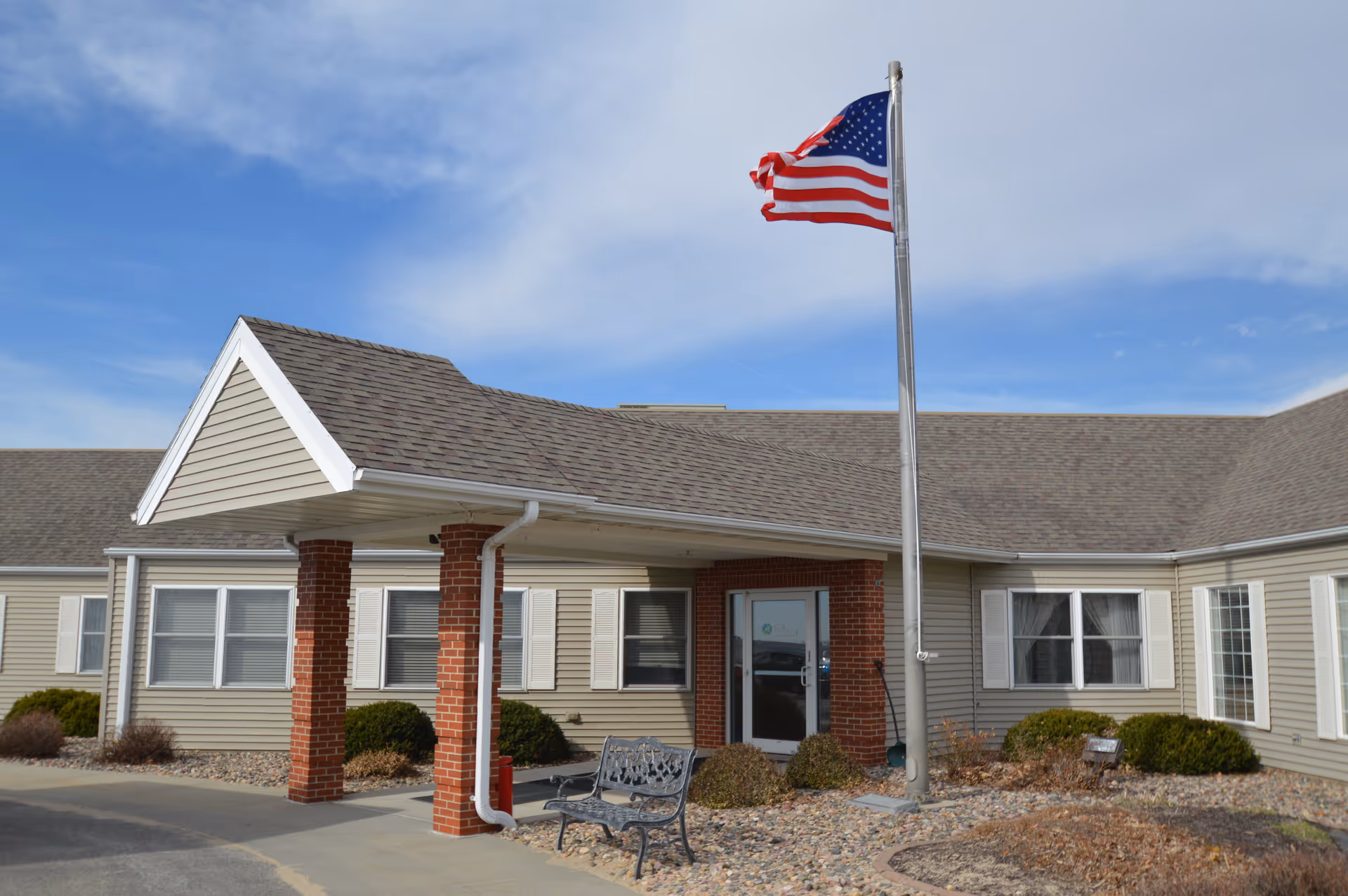 Front entrance of a single-story care facility with a covered portico, brick columns, a bench, and an American flag on a flagpole.