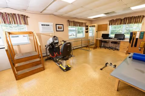 A physical therapy or rehabilitation room with exercise equipment including a recumbent bike, wooden stairs for climbing exercises, parallel bars for walking support, a treatment table, and two computer workstations along the far wall. The room has beige walls, windows with striped valances, and a cork or linoleum floor.