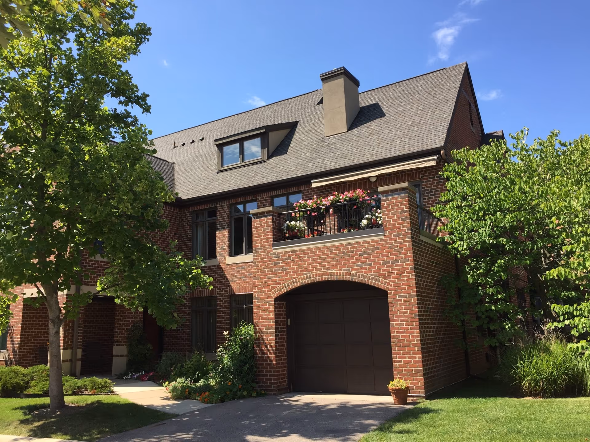 Front view of a two-story red brick townhouse with a garage, a flower-filled balcony, and trees on a sunny day.