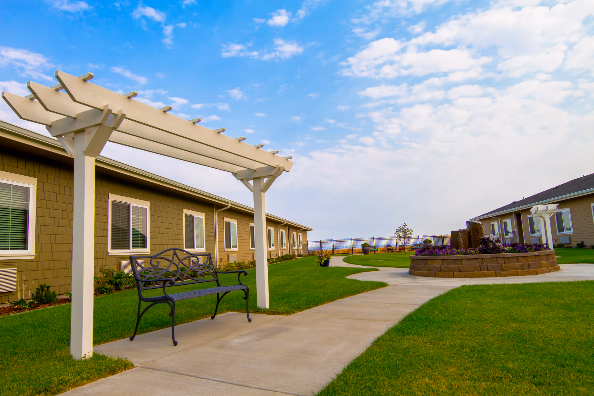 Outdoor courtyard of a memory care facility with a white pergola and bench beside a paved path, lawn, and a circular planter/fountain between single-story buildings.