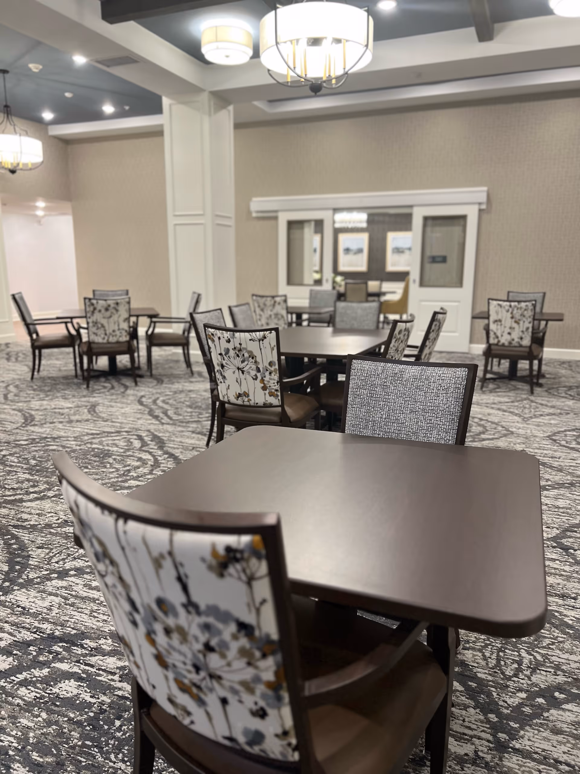 Spacious communal dining area with square dark wood tables and floral-patterned chairs under chandeliers.