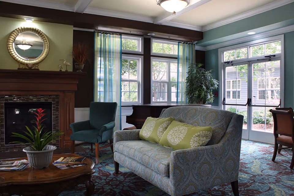 A cozy living room area in an assisted living facility featuring a patterned loveseat with three green decorative pillows, a green armchair, a wooden coffee table with a potted plant and magazines, a fireplace with a round mirror above it, large windows with sheer blue curtains, a piano in the corner, and a glass door leading outside.