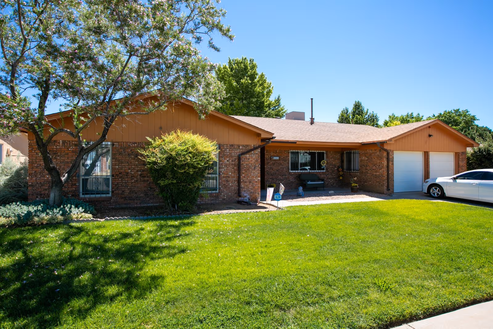 Single-story brick house with a brown roof and two white garage doors, surrounded by green grass, trees, and shrubs under a clear blue sky. A white car is parked in the driveway.