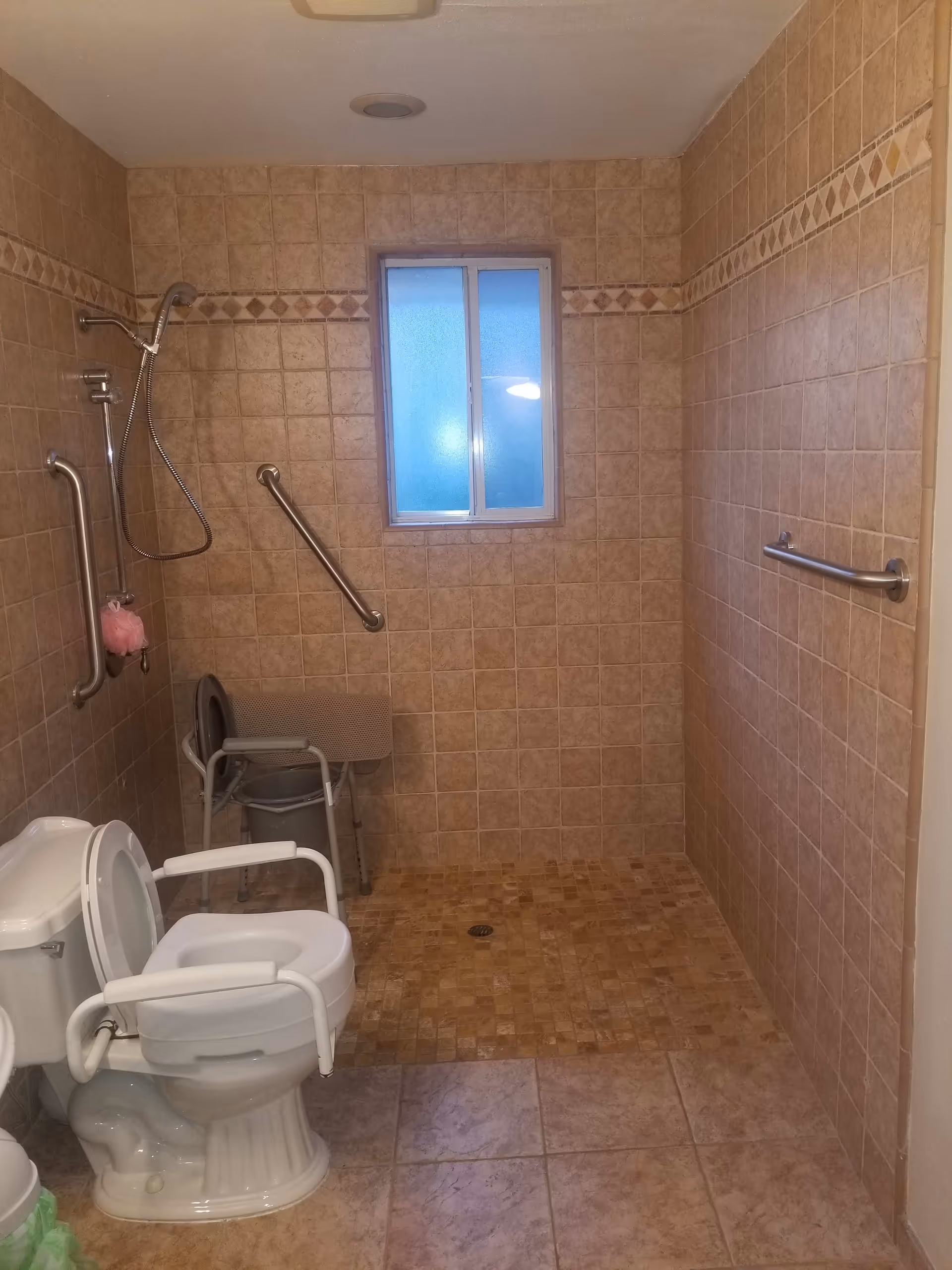 A bathroom with beige tiled walls and floor, featuring a toilet with white safety rails, a shower area with a handheld showerhead, grab bars on the walls, a small window with frosted glass, and a shower chair.