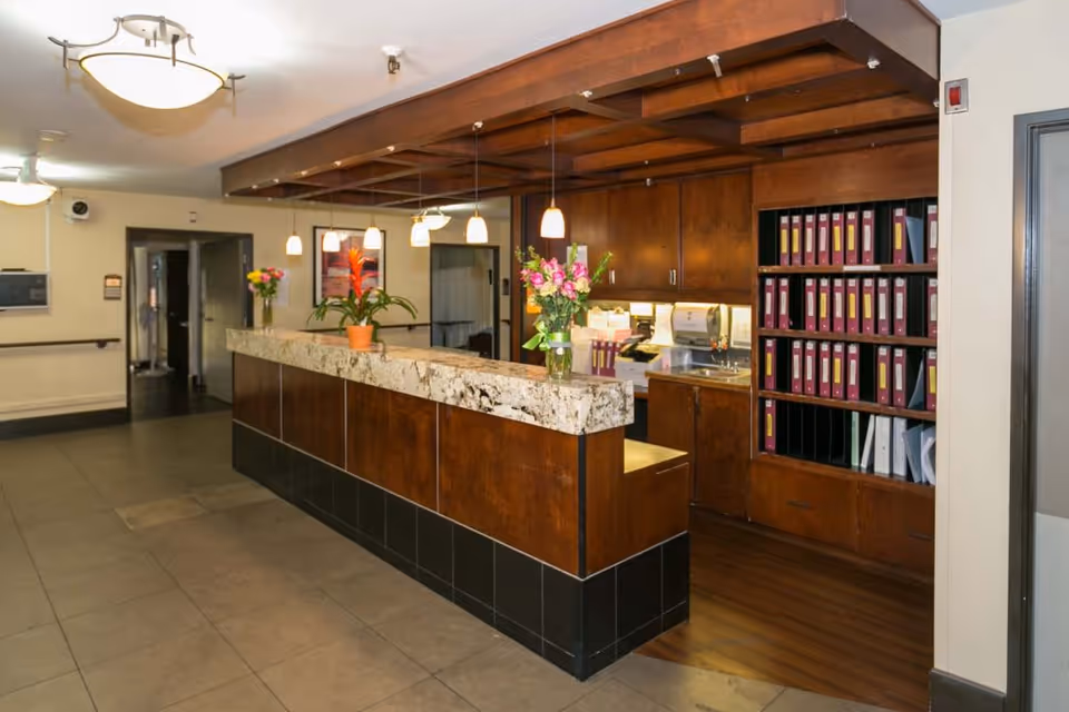 Reception area with a long marble countertop and wooden paneling. There are several hanging pendant lights above the counter and flower arrangements placed on it. Behind the counter are wooden cabinets and shelves filled with organized binders. The floor is tiled, and there is a hallway visible in the background.
