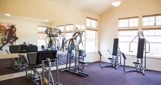 A bright fitness room with exercise equipment including a treadmill, elliptical machine, and weight machines. The room has large windows letting in natural light and a wall mirror reflecting the equipment.