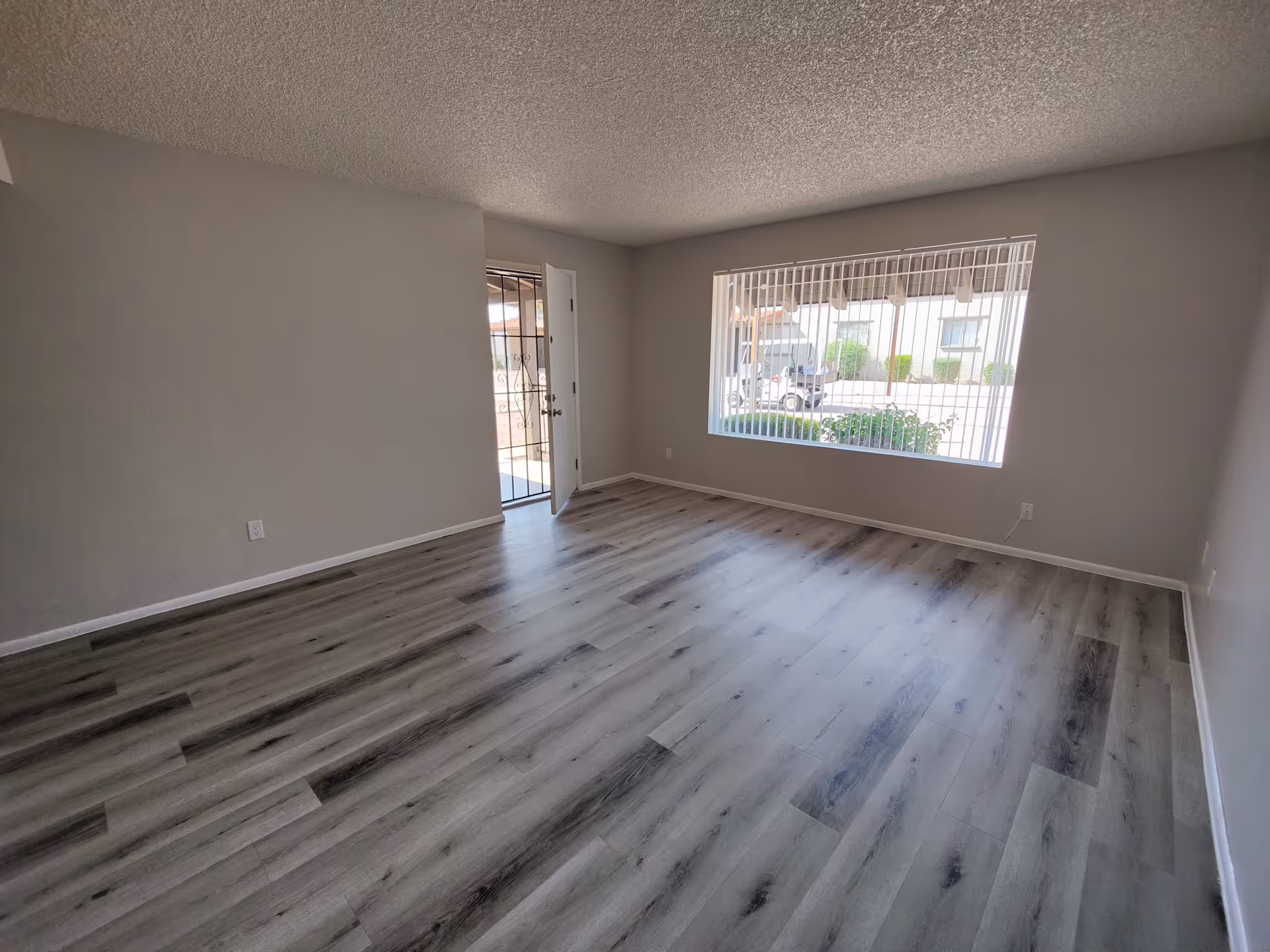 Empty room with light gray walls and wood-patterned vinyl flooring. A large window with vertical blinds looks out onto a parking area with a golf cart and some greenery. A door with a security screen is open, leading outside.