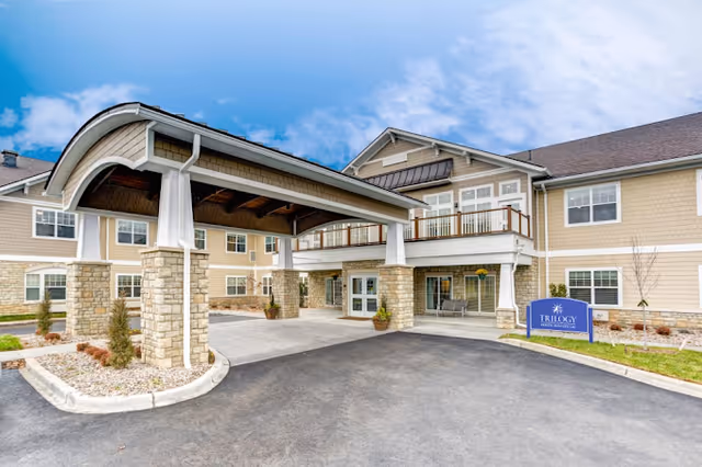 Exterior view of a senior living facility with a covered entrance supported by stone pillars, beige siding, multiple windows, and a sign that reads 'Trilogy Senior Living'. The sky is partly cloudy.