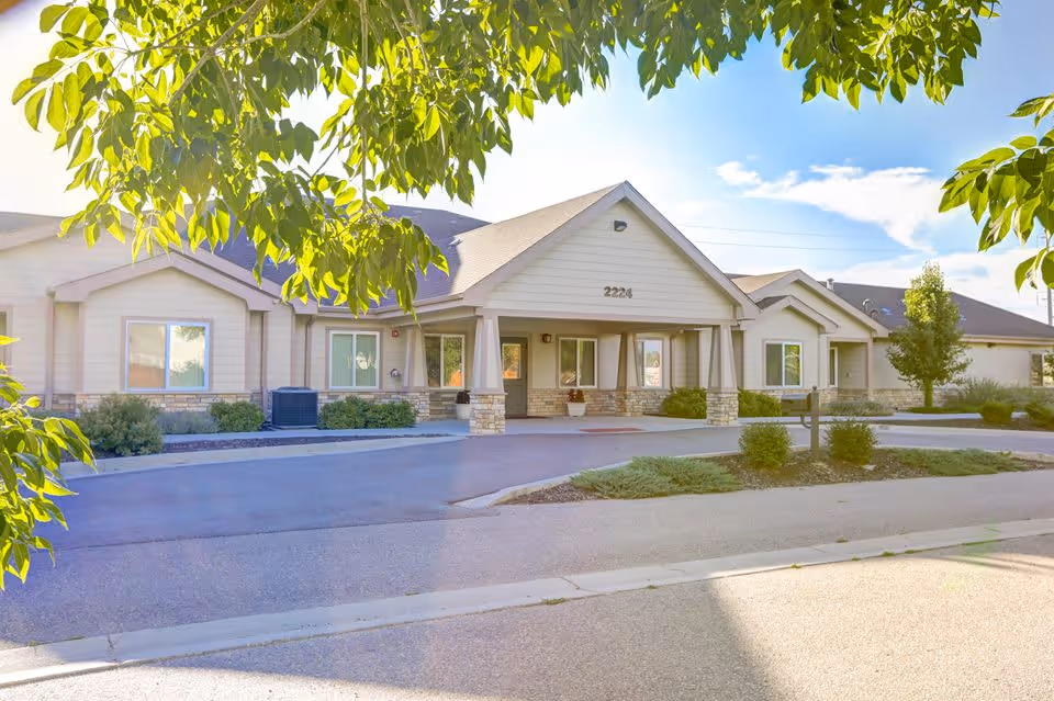 Exterior view of a single-story senior living facility building with beige siding and stone accents, a covered entrance with pillars, surrounded by greenery and trees under a bright blue sky.