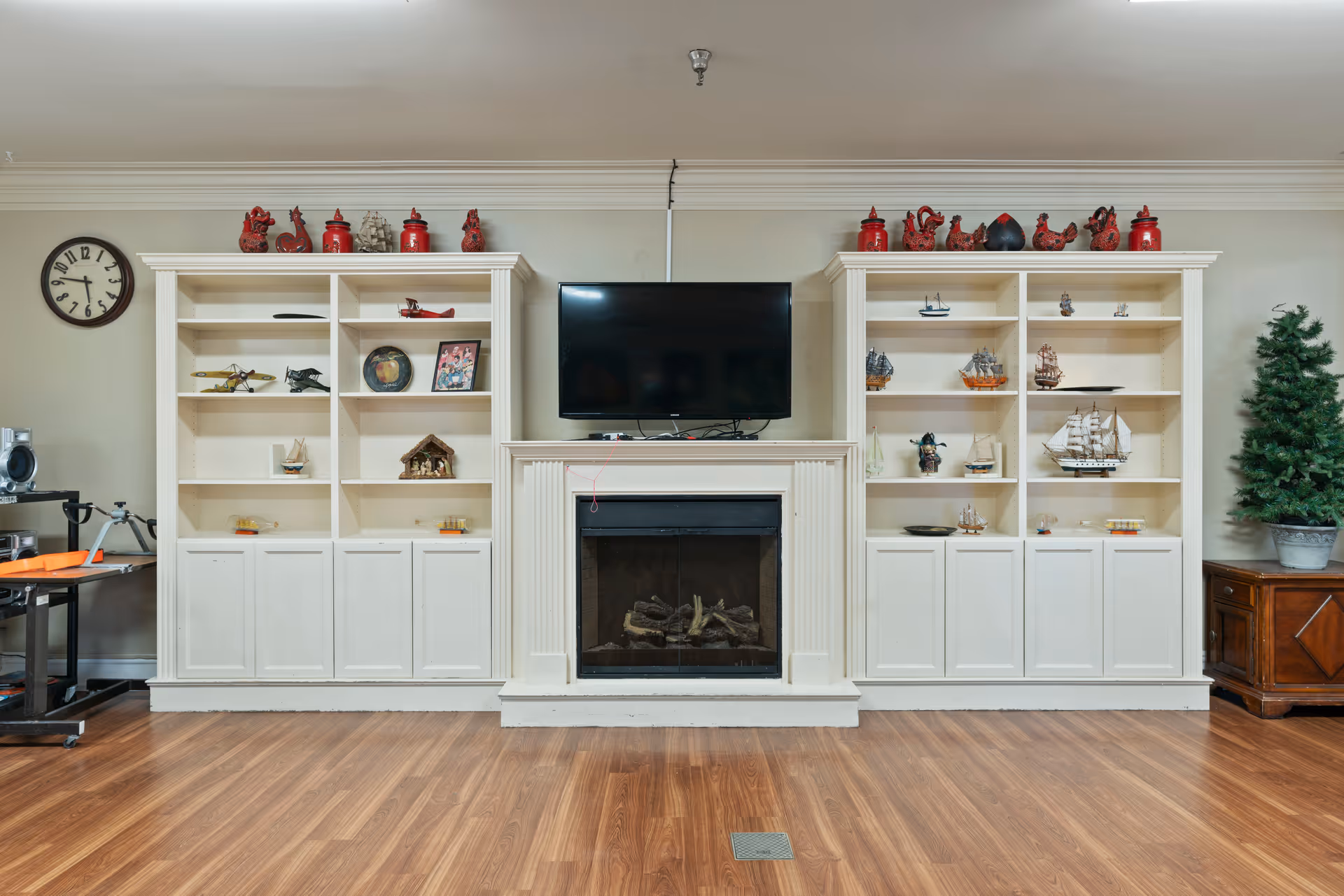 A cozy interior room featuring a white fireplace with a flat-screen TV mounted above it, flanked by two white built-in bookshelves filled with decorative items including model ships and red ceramic pieces. To the right is a small green potted tree on a wooden cabinet, and to the left is a clock on the wall and some equipment on a table. The floor is wooden with a vent in the center.