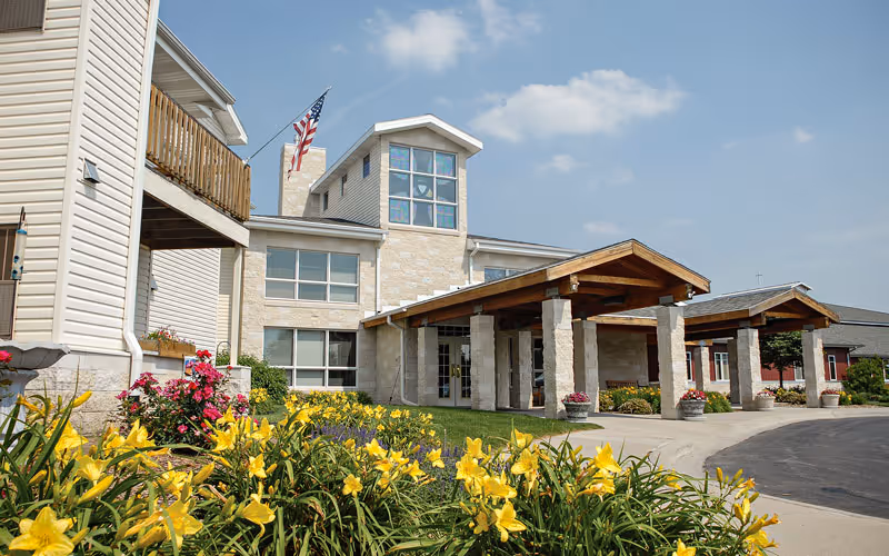 Exterior view of Immanuel: Trinity Village building with a covered entrance supported by stone pillars, surrounded by a garden with yellow flowers and a clear blue sky.