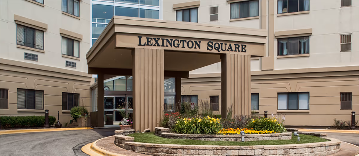 Entrance to a senior living facility named Lexington Square with a covered drop-off area, surrounded by a circular flower bed with yellow and orange flowers, and the building in the background.
