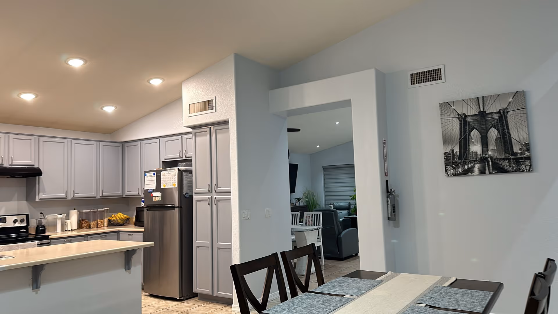 Interior view of a modern kitchen and dining area in an assisted living facility. The kitchen features light gray cabinets, a stainless steel refrigerator, a stove, and a countertop with various kitchen items including jars and a bowl of bananas. The dining area has a dark wooden table with chairs and a table runner. A black and white photo of a bridge is hung on the wall. In the background, a living room area with seating is partially visible through an open doorway.
