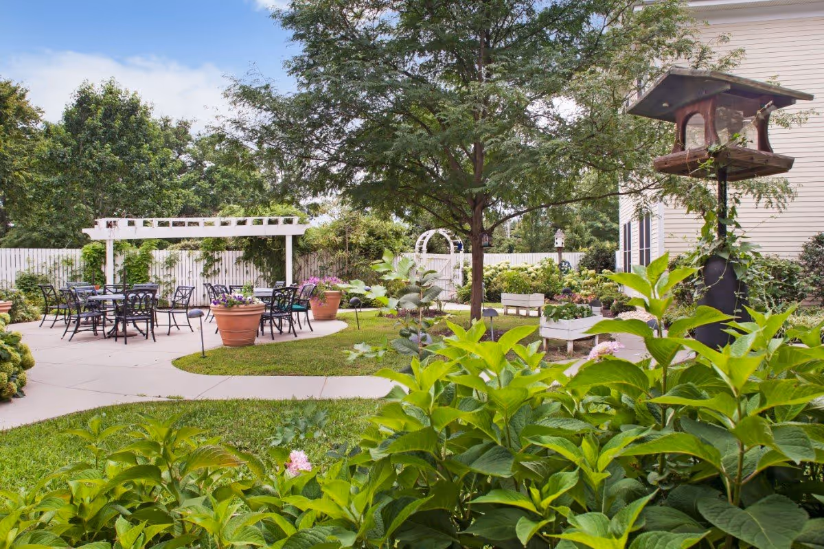 A peaceful outdoor garden area with green plants, trees, and flowers. There are several black metal tables and chairs arranged on a paved patio under a white pergola. A bird feeder is mounted on a post near the right side of the image, and a white fence encloses the garden space.
