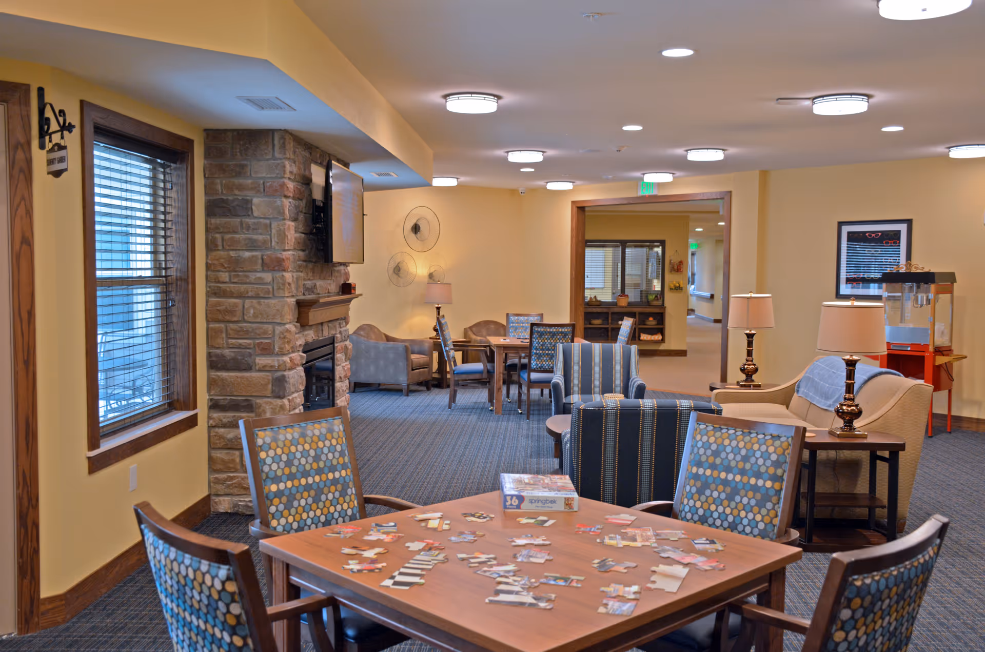 A cozy common area in a senior living facility featuring a table with a partially completed jigsaw puzzle and four patterned chairs. The room has a stone fireplace with a mounted TV above it, several armchairs and sofas, lamps, and warm yellow walls. There is a popcorn machine in the background and a framed picture on the wall. Large windows with blinds allow natural light into the space.