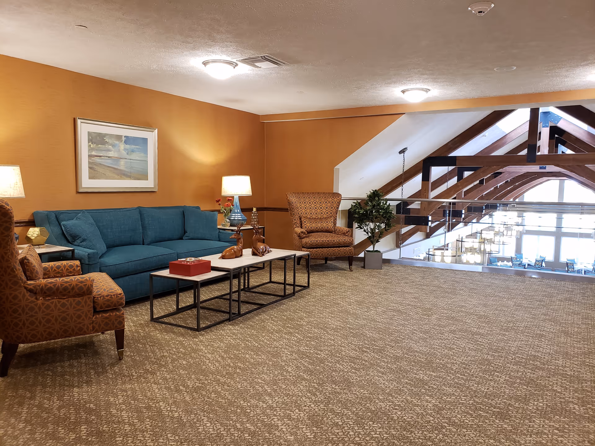 A cozy sitting area in a retirement community with a teal sofa, two patterned armchairs, a set of three nested coffee tables with decorative items, two table lamps, a potted plant, and a framed beach painting on an orange wall. The area overlooks a large open space with wooden beams and large windows letting in natural light.