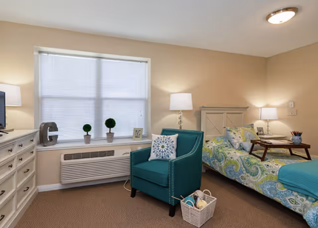 A cozy furnished bedroom with a bed, teal armchair, dresser, window, and lamps.