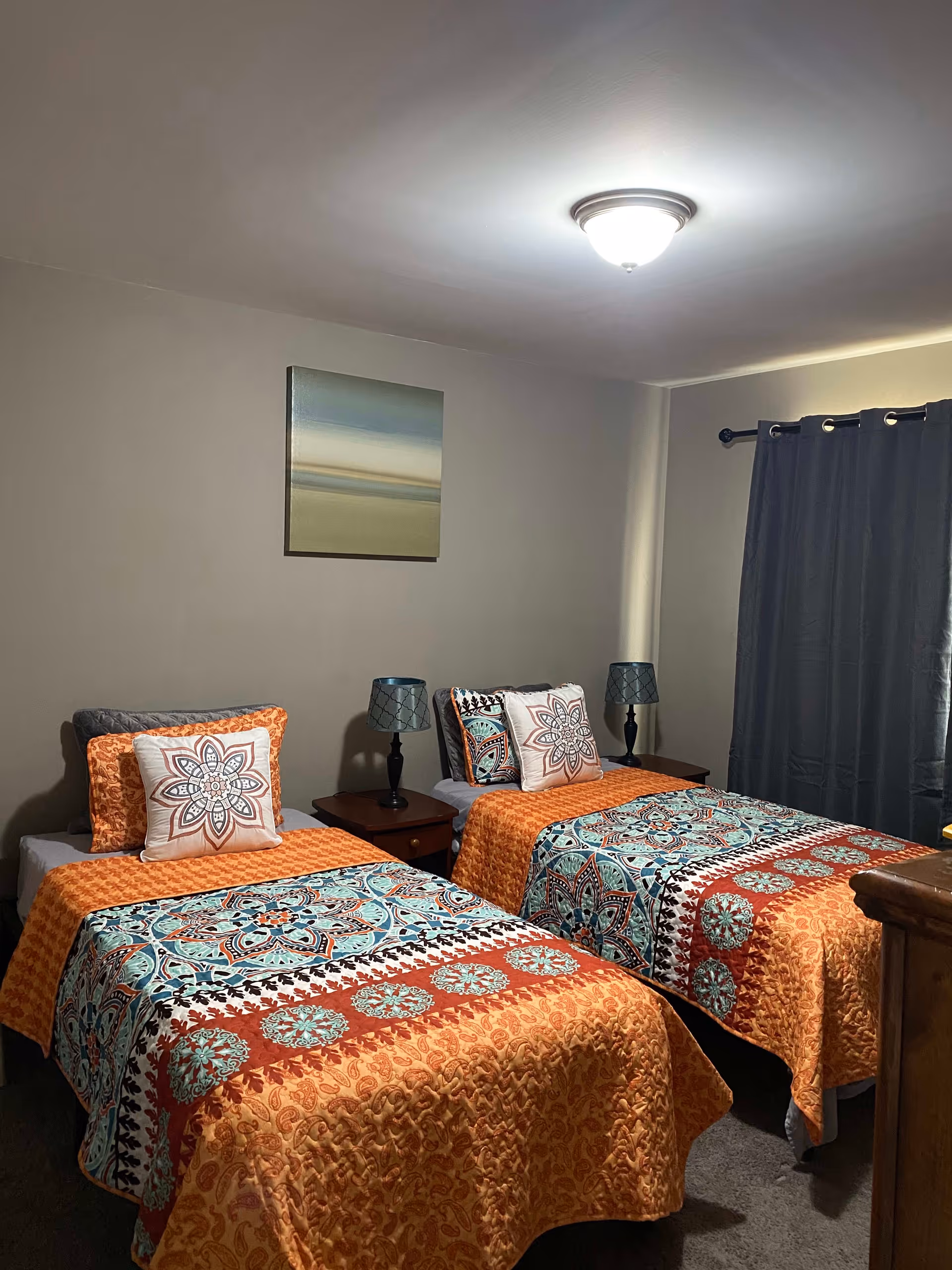 A bedroom with two twin beds side by side, each with colorful orange, blue, and red patterned bedspreads and matching pillows. Between the beds is a small wooden nightstand with a black lamp. A simple abstract painting hangs on the beige wall above the beds. A window with dark curtains is visible on the right side of the room.