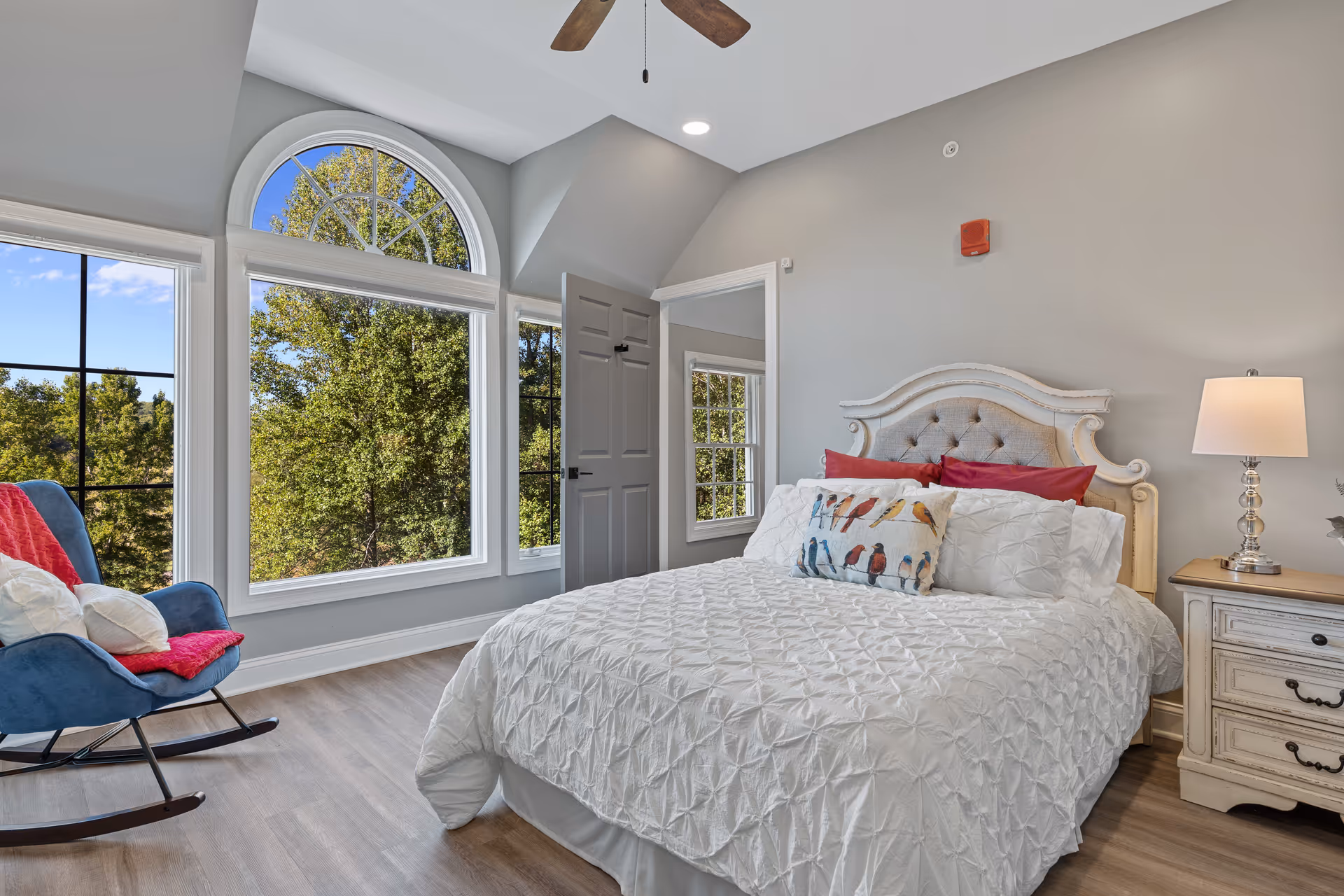 Bright bedroom featuring a tufted headboard bed, nightstand with a lamp, a rocking chair and large arched windows overlooking trees.