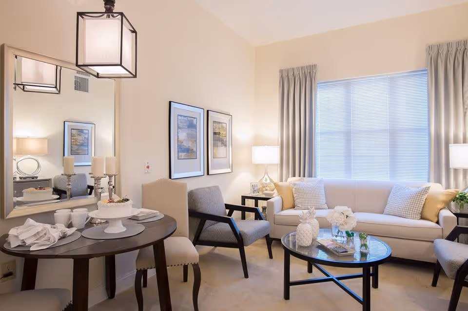 A bright and cozy living room area with a beige sofa adorned with yellow and checkered pillows, two gray armchairs, a round glass coffee table with decorative items, and a small round dining table with chairs. The room features a large window with blinds and curtains, two framed pictures on the wall, and soft lighting from table lamps and a hanging light fixture.
