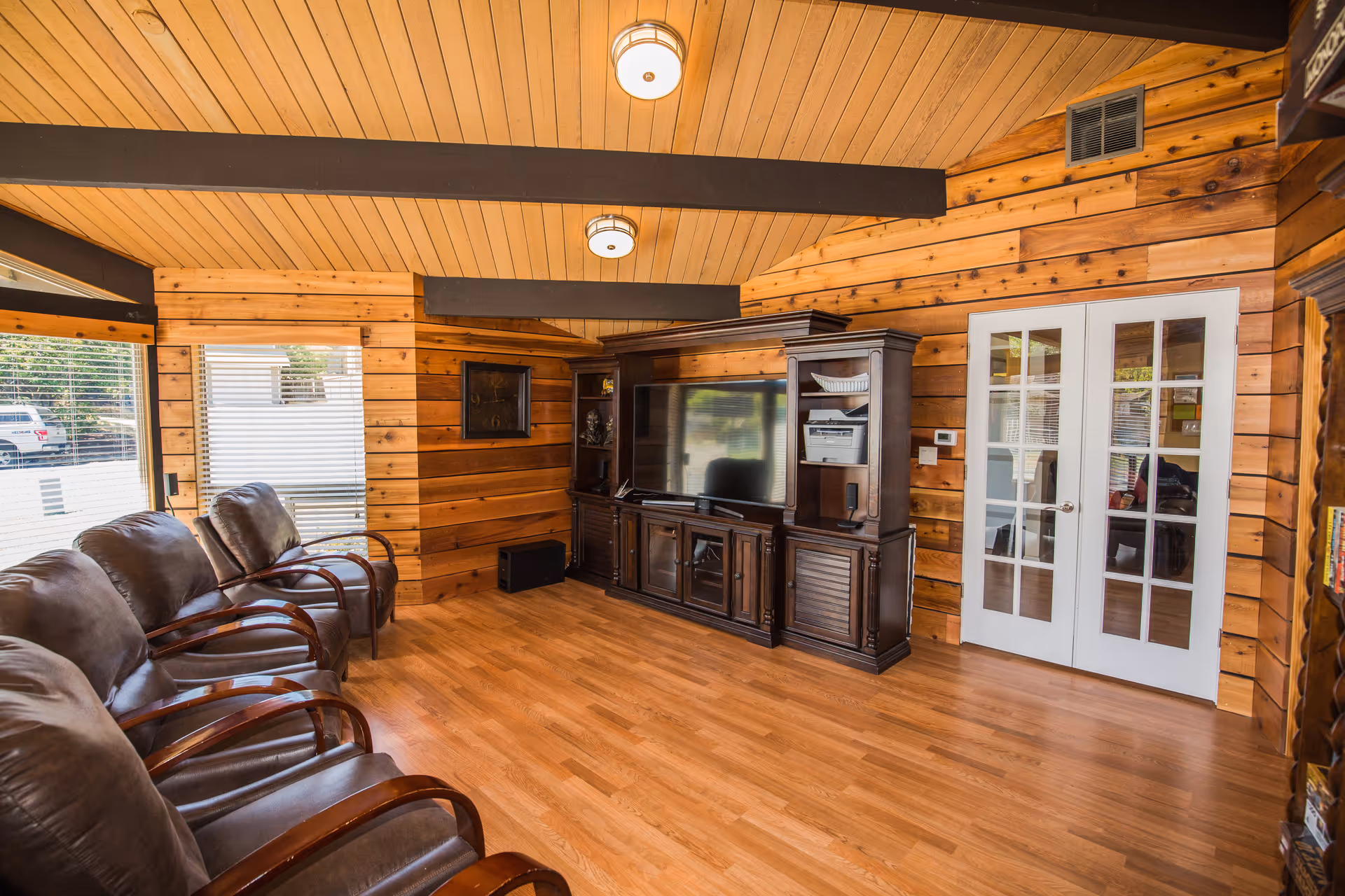 A cozy living room with wooden walls and ceiling, featuring a row of brown leather chairs with wooden armrests along one side. A large dark wood entertainment center with a flat-screen TV and shelves is positioned against the wall. There are two white French doors with glass panes on the right side, and windows with blinds on the left side letting in natural light.