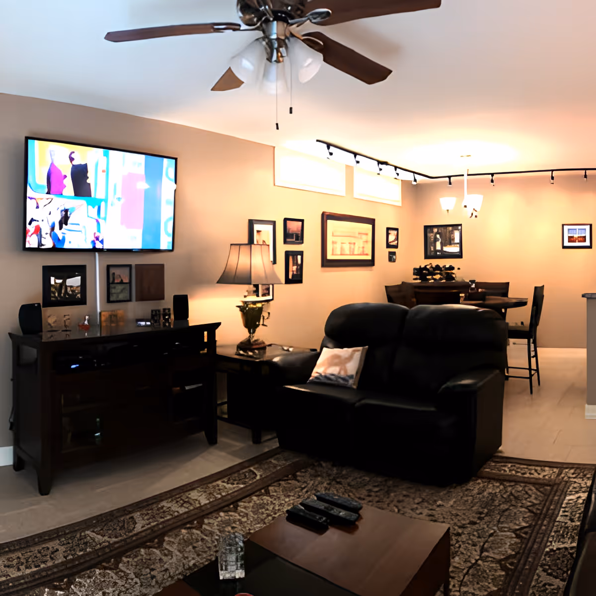 A cozy living room with a black leather loveseat, a wooden coffee table with remote controls, a patterned area rug, a wooden TV stand with a flat-screen TV mounted above it, a table lamp, and framed pictures on the beige walls. In the background, there is a dining area with a round table and chairs, illuminated by ceiling lights and track lighting.