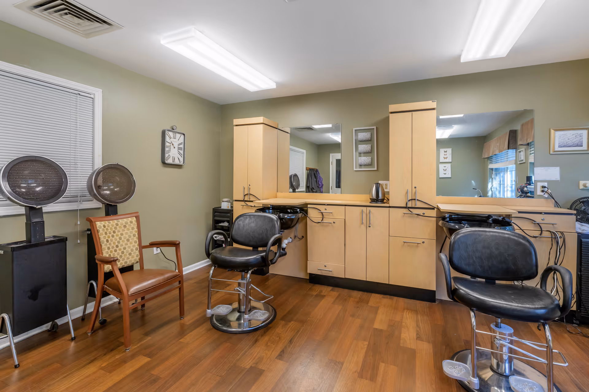 Interior view of a hair salon area with two black salon chairs in front of a large mirror and wooden cabinets. There are two hair dryers on stands near a window with closed blinds, a wooden chair with patterned upholstery, and a clock on the green wall. The floor is wood, and the ceiling has fluorescent lights.